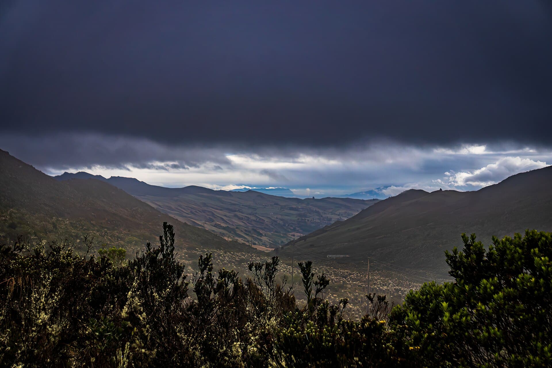 Valle de alta montaña con arbustos de páramo en primer plano, laderas con parcelas al fondo y un cielo cargado de nubes oscuras, en el complejo de páramo de Sumapaz